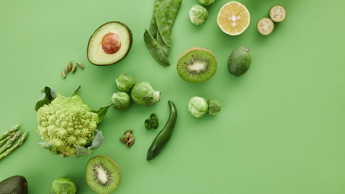 Top-down view of assorted fresh green vegetables and fruits on a vibrant green background.